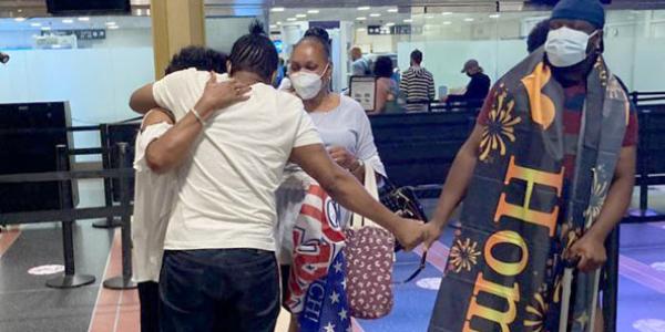 Photo of a man in a white shirt and black pants, back to the camera, hugging two family members at the airport. His arm is outstretched to his right and he is holding the hand of another family member, who has a banner with wrapped around his shoulders, with the word "Home" visible in large letters.