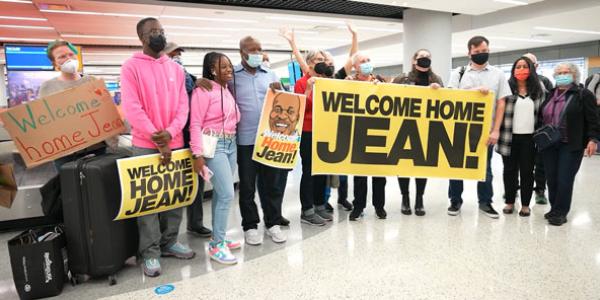 Photo of Jean in the middle of a large group of family and friends who have met him at an airport baggage claim area to welcome him home. People hold a banner and three different sights with the message "Welcome Home Jean!"