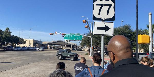 Photo of a group of people walking down the street in Brownsville Texas, under a clear blue sky, toward the a port of entry at the U.S. border with Mexico.
