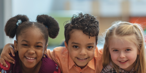 A photo of three cute children smiling leaning over a table and smiling. The child in the middle has their arms around the child on each side of them.