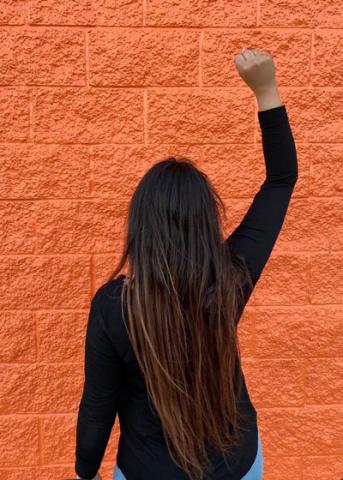 A photo of a woman with long dark hair, wearing a black shirt and with her back turned to the camera and her right fist in the air, with an orange stone wall in front of her.