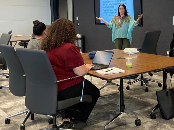 One professional woman at the front of a conference room presenting enthusiastically with slide deck behind her. Two professionals are sitting at a table and listening.