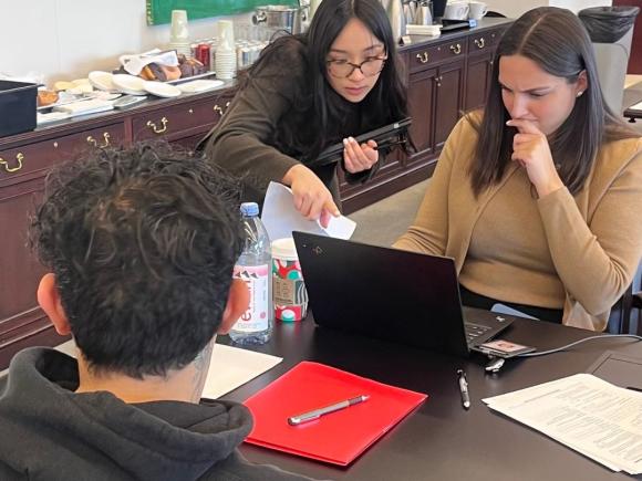 A woman attorney sitting at a desk across from her young male client. She is looking at her laptop, with another woman peering over her shoulder.