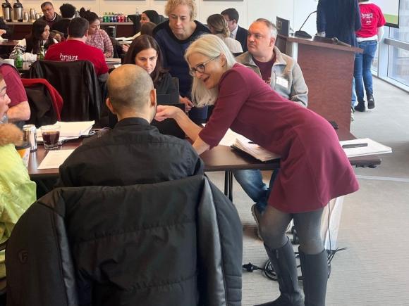  A person in a red dress assists another person with paperwork at a busy clinic. Other participants are engaged in conversations in the background, decorated with colorful artwork.