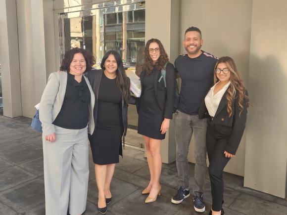 A woman stands outside a courthouse next to two woman attorneys and a couple of her friends. All are smiling because she just won asylum.