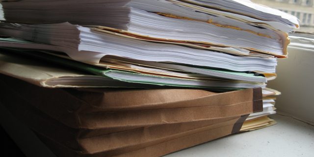 A brown accordion file and manila folders filled with papers stacked on a ledge next to a window that looks out onto a city building.