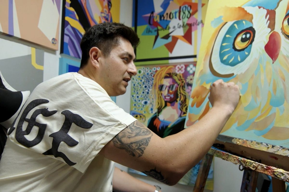 Man painting. A man sits at an artist easel working on a painting of an owl. In the background an assortment of bright-colored paintings decorate the wall.
