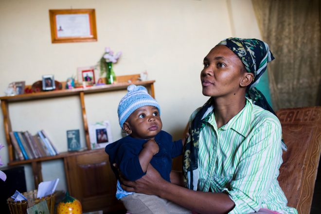 Photo of a woman wearing a head scarf, sitting on a couch with her baby son on her lap, looking at something out of frame to the upper left.