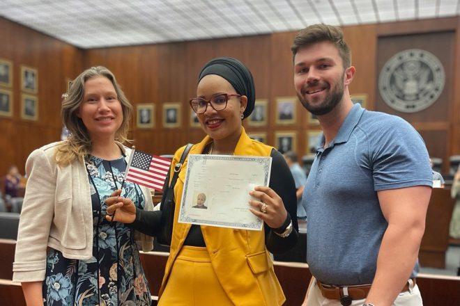 Photo of a woman holding a small U.S. flag and a certificate of U.S. citizenship. She stands between the two attorneys who represented her in her naturalization case.