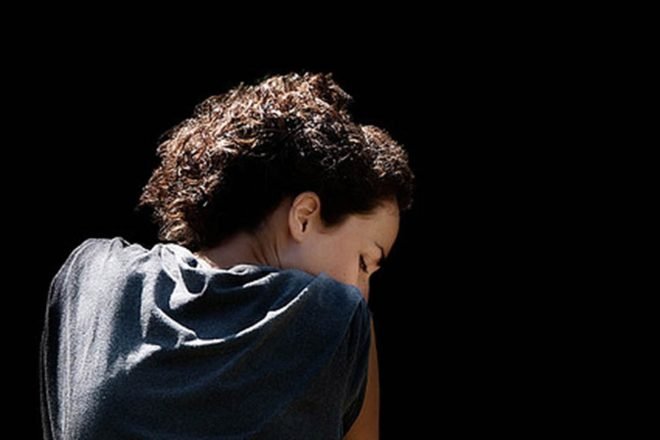 Photo from behind of the shoulders and profile of a woman with curly hair, looking to the right and down with a pensive expression.