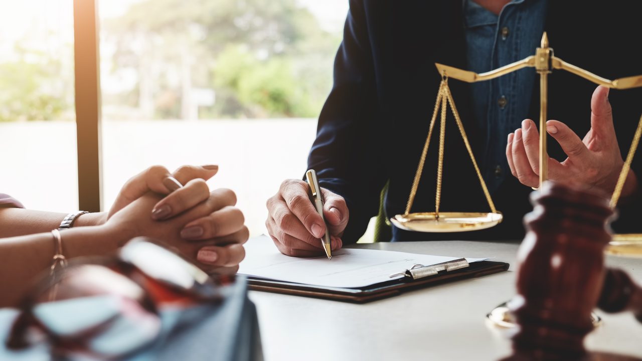 Image of a client's hands clasped and their attorney's hands writing in a notepad. In the foreground is a gavel and justice scales.