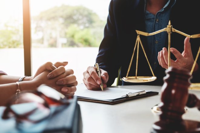 Image of a client's hands clasped and their attorney's hands writing in a notepad. In the foreground is a gavel and justice scales.