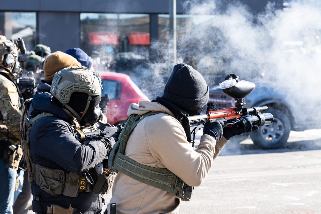 Masked ICE agents shoot rubber bullets with smoke and teargas visible in background in Minneapolis. Credit Chad Davis CC BY 4.0